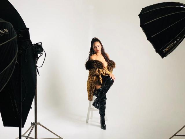 VenusChaud with curly dark hair, wearing a brown fur coat off-shoulder, black thigh-high boots, and no underwear, sitting on a white stool between two black studio lights.
