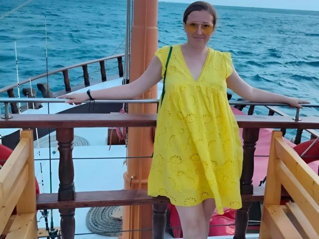 AngellaSexy stands on a boat wearing a yellow dress with small holes, yellow sunglasses, and a black wristband. Her arms rest on a wooden railing, ocean behind.