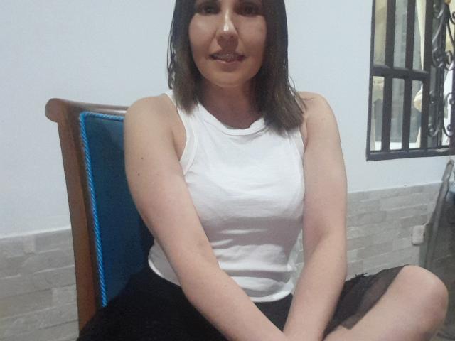AdelaShery, avec des cheveux bruns mi-longs, un haut blanc sans manches, une jupe noire, assise en tailleur, sur une chaise rembourrée bleue, les bras reposant sur ses genoux, souriant légèrement, avec une poitrine de taille moyenne, et une peau claire..