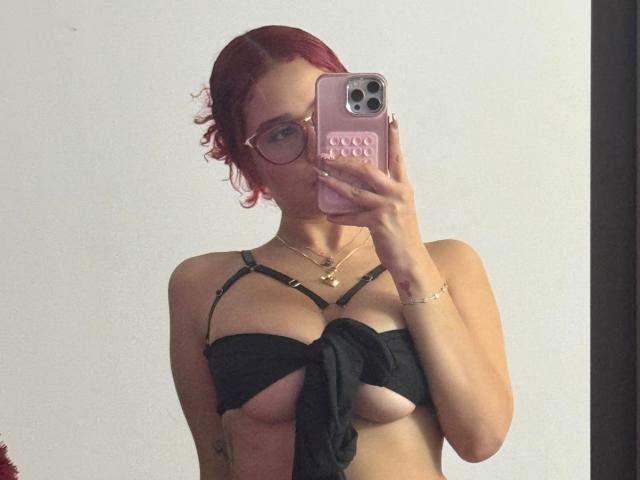 ValeryScot with red hair, glasses, and a black strappy top that exposes her small tits and nipples, taking a mirror selfie with a pink phone.