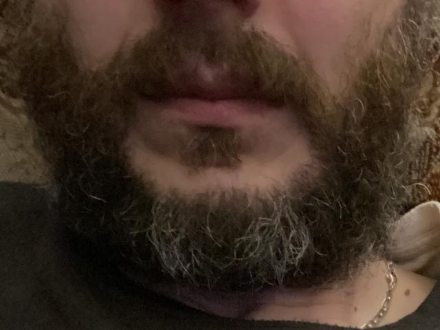 Il volto di Semoris, con una folta barba e baffi castano ricci, labbra leggermente schiuse di colore rosato, e una visibile collana d'oro nell'angolo in basso a destra. L'immagine è sfocata..