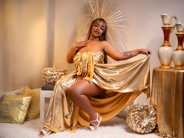 MadinsonLopez with golden dress, showing her ass, holding fabric, wearing white strappy heels, headpiece with rays, tattoos on arm, sitting on white rug, surrounded by gold decor.