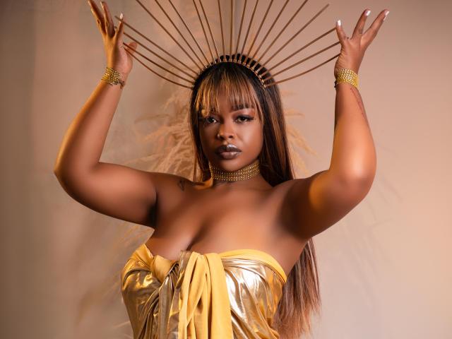 MadinsonLopez with dark skin, large tits, wearing a shiny gold strapless dress, gold headpiece, and feathered halo, arms raised, gold bracelets, and choker, looking confident.