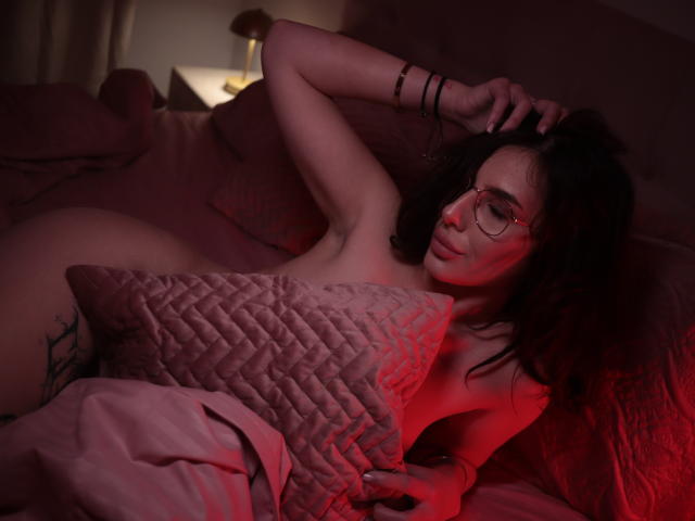 EvaBrianna, naked, with glasses, lying on a bed, holding a pink quilted pillow over her tits, red light, arm raised, wrist bracelet, dark curly hair, tattoo on left hip.