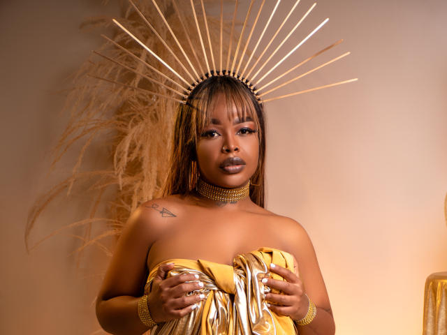 MadinsonLopez with golden headpiece, holding shiny gold cloth over her tits, wearing gold jewelry, and looking at the camera with a neutral expression.