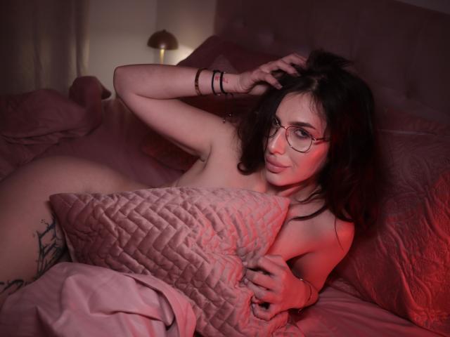 Nude EvaBrianna with glasses, holding a pink quilted pillow, covering her tits, red bedroom light, tattoo on left hip, arm bracelet, looking at camera, lying on pink bed.