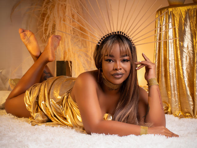 MadinsonLopez is lying on a white rug, wearing a shiny gold dress and headpiece, with her ass up and legs crossed. She's looking at the camera, smiling.