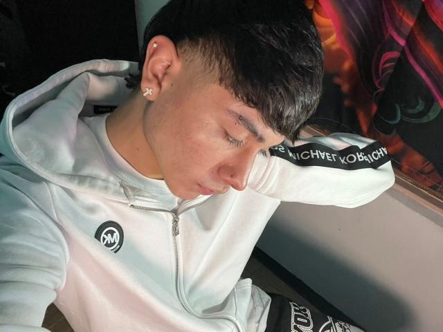 ImJuanda with short dark hair, white hoodie, small ear stud, nose ring, eyes closed, head down, left hand behind head, sitting, dark hair, white hoodie, dim light.