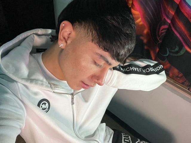 ImJuanda with short dark hair, ear studs, white hoodie, and black arm band, eyes closed, head tilted down, pink lips, slight nose piercing, white earbuds, and relaxed expression.