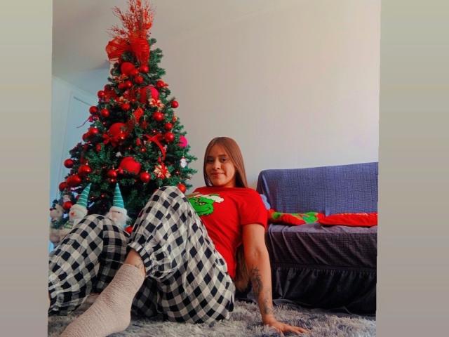 BelletinaXx with brown hair, red shirt, and checkered pants, sitting on a gray rug, legs up, showing white socks, tattoo on right arm, Christmas tree in the background.