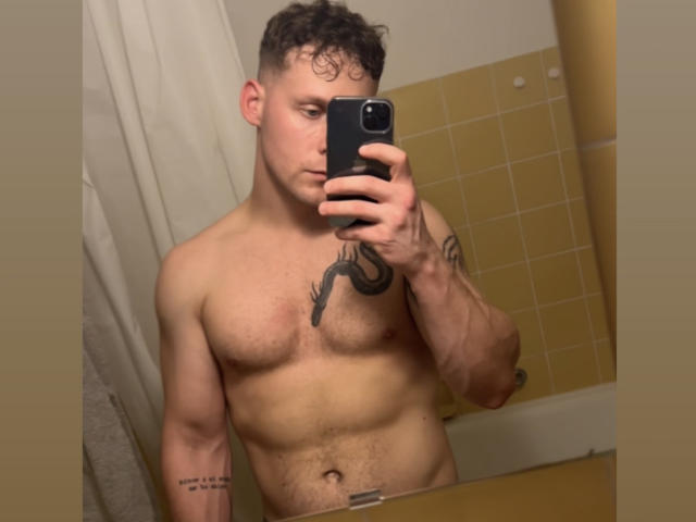 AdamBuran, shirtless with curly brown hair, holding a phone in front of his face, showing a tattoo on his right chest and another tattoo on his left forearm.