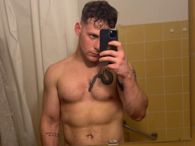 AdamBuran, shirtless, muscular, with a snake tattoo on his chest, holding a phone, taking a mirror selfie, showing a serious expression, in a bathroom with yellow tiles.