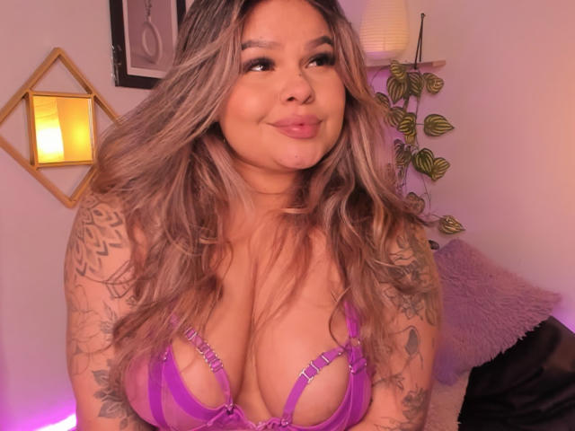 JulianaSoussa, con lunghi capelli biondi ondulati, indossa un reggiseno viola con spalline sottili, che mette in mostra il suo seno grande e prosperoso, e ha tatuaggi sulle braccia, sorridendo dolcemente alla fotocamera..