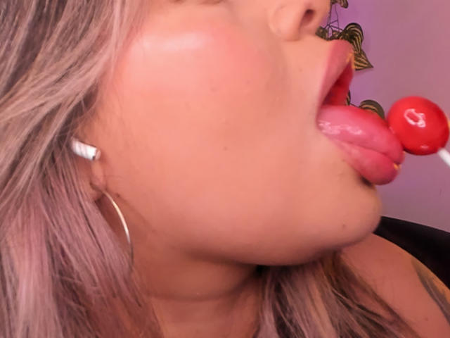 JulianaSoussa's pink-lipped mouth is around a red lollipop, her tongue out, with her ass in the background and white earbuds in her pinkish-brown hair.