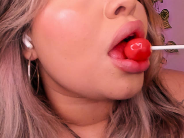 JulianaSoussa's face, lips open with a red heart-shaped lollipop in her mouth, pinkish blonde hair, white earbuds, and gold hoop earrings, slight blush on her cheek.