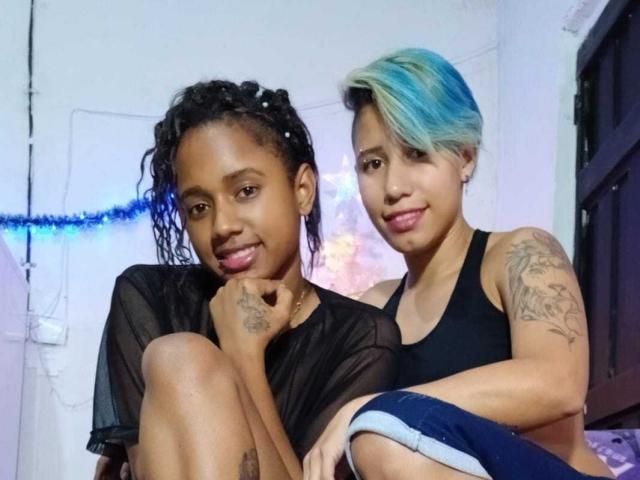 ElectrayAlondra with curly black hair and black sheer top, and ElectrayAlondra with blue-green hair, black tank top, and tattooed arm, both sticking out tongues, sitting together.