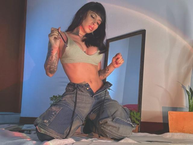 LalaVega ajoelhada na cama, vestindo uma blusa cropped verde, jeans abertos, tatuagens nos braços, mãos levantadas, cabelo preto, batom vermelho, sexy, atraente, abdômen visível, luz suave, espelho ao fundo..