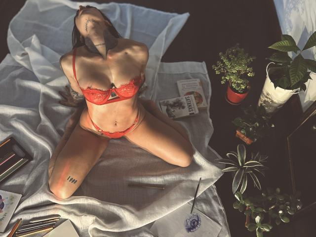 LalaVega with red lace bra and panties, sitting on white cloth, arms behind head, tattooed, tattoo on neck, sunlight, plants, book, pencils, and notebook on dark floor.