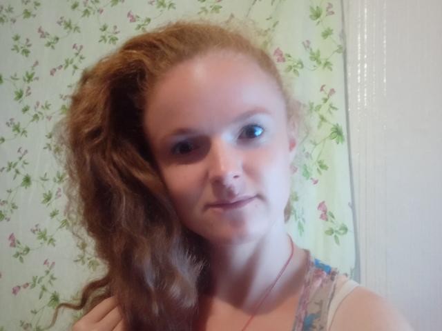 RaeJeannie has curly red hair, blue eyes, and a slight smile. She's wearing a colorful top, with a red necklace, and a subtle, cute, attractive face.