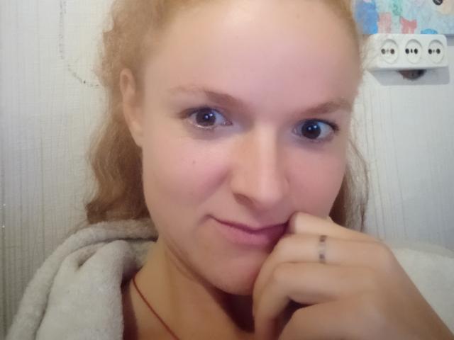 RaeJeannie has pale skin, blue eyes, and blonde hair. She's smiling with her hand on her chin, wearing a gray hoodie. Her ring finger has a small tattoo.