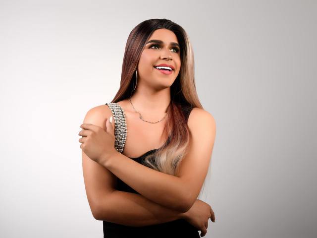 NathalyHotty with long brown-to-blonde ombre hair, black sleeveless top, and chain necklace, smiling, arms crossed, showing small tits, and pink tongue slightly out.