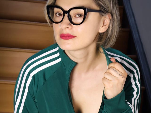 KatParadise, avec des lunettes, une veste verte et du rouge à lèvres, montre sa poitrine en posant une main sur elle, a des tatouages et un piercing au nez, et se tient sur des marches en bois..