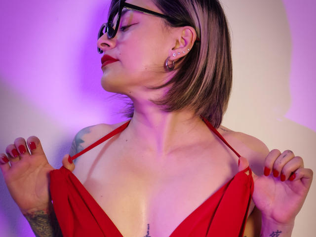 KatParadise, con cabello castaño corto, gafas, lápiz labial rojo, piercing en la nariz y aretes, sosteniendo las tiras de un vestido rojo, mostrando pequeños tatuajes y uñas pintadas de rojo, con iluminación púrpura..