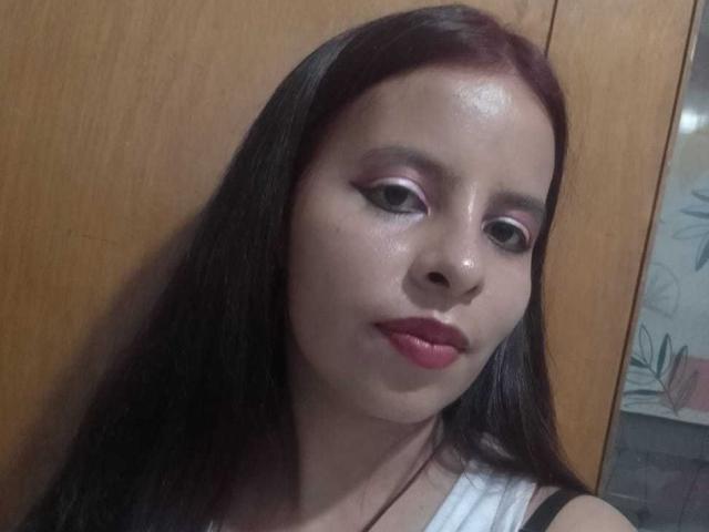 JuliaWaine has shiny, pale skin, dark straight hair, pink eyeshadow, black eyeliner, red lipstick, and a neutral expression. She's wearing a white top with black straps.
