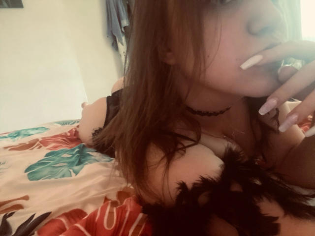 Toffee69 met lang bruin haar, een zwarte choker en roze nagels, liggend op een bed met bloemenprint lakens, met haar hand in de buurt van haar mond, waarbij haar grote borsten en een zwarte veerpendant zichtbaar zijn..