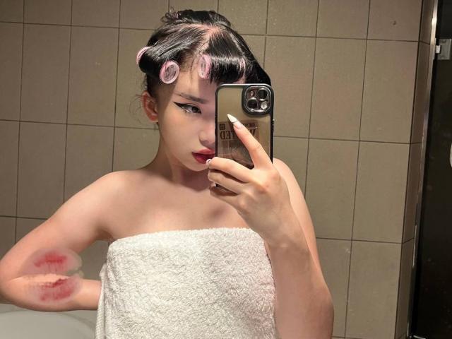 EveLovelace con i bigodini rosa, rossetto scuro, un asciugamano bianco che copre il seno, un livido rosso sul braccio sinistro, che si fa un selfie allo specchio con un iPhone nel bagno..