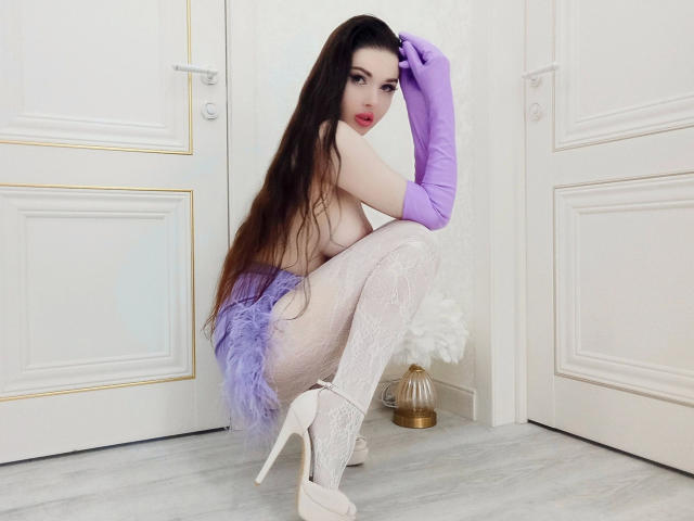 KatrinaBonita-hot, squatting, with long brown hair, purple gloves, white lace tights, and high heels, wearing a purple feather skirt, showing her tits and pale skin, in a room.