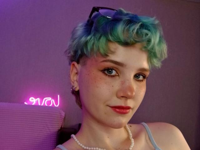 ShellyLove69 has short, curly blue-green hair, freckles, and wears glasses on her head. She's smiling with red lipstick, a pearl necklace, and a neon pink sign in the background.