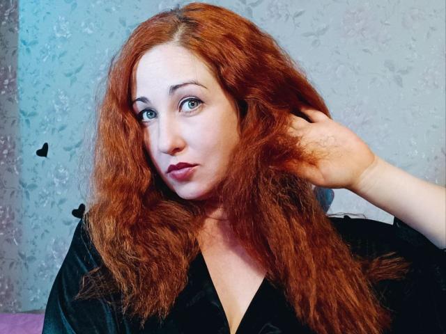 JillSilvies with red curly hair, pale skin, and blue eyes, wearing a black shirt, pouting with a hand in her hair, looking at the camera. Her tits are visible.