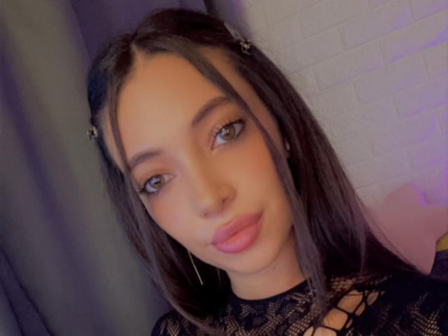 LaylaKrista with long brown hair, braided, wearing black lace top, pink lips, hoop earrings, and blue eyes, looking at camera with slight smile, medium skin tone.