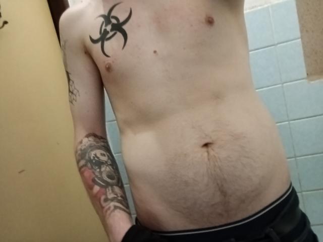 KuroshHype's shirtless torso with a biohazard tattoo on the left chest, a tattooed arm, and a hairy belly, wearing black pants, in a bathroom.