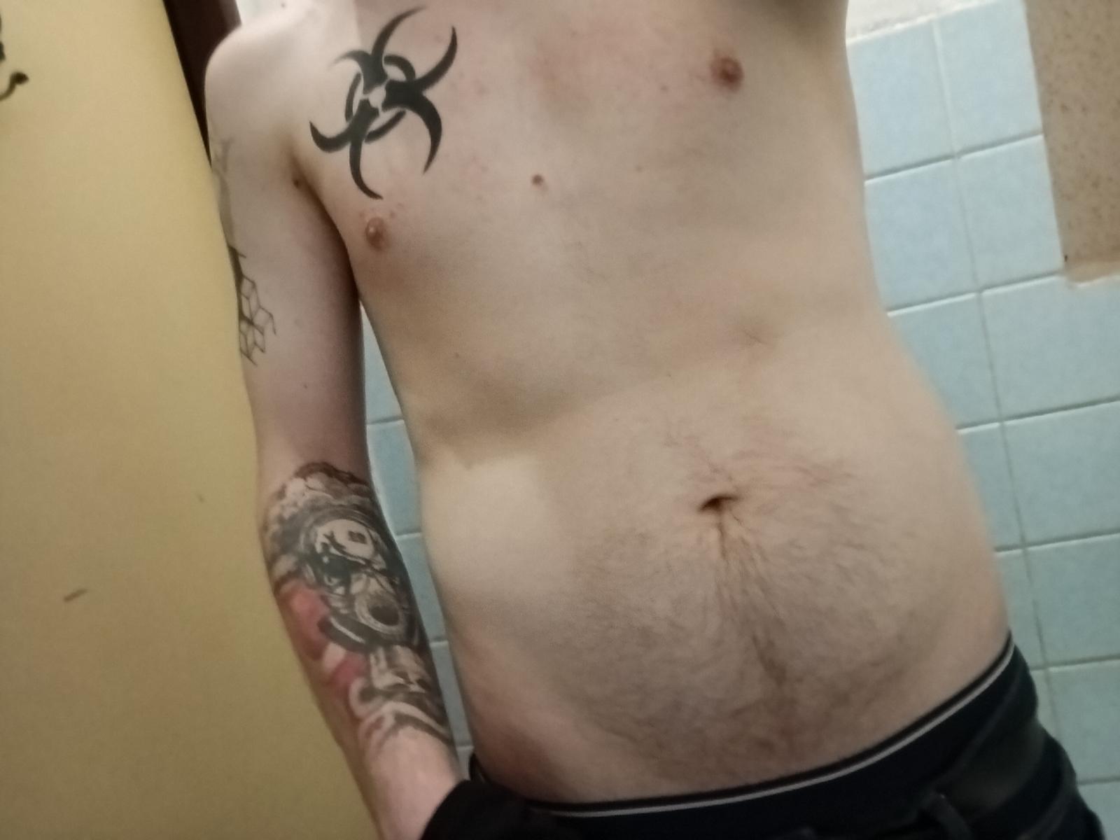 KuroshHype's shirtless torso with a biohazard tattoo on the left chest, a tattooed arm, and a hairy belly, wearing black pants, in a bathroom.