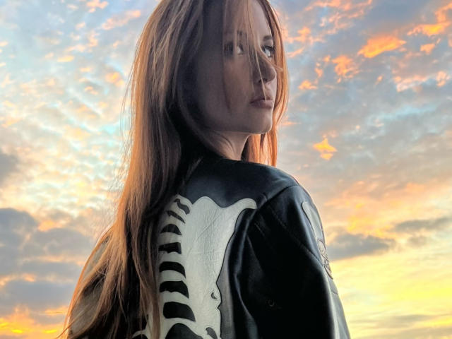 TheTyQueen with long, straight brown hair wearing a black leather jacket with white skeleton print on the back, looking over her shoulder at a sunset sky.