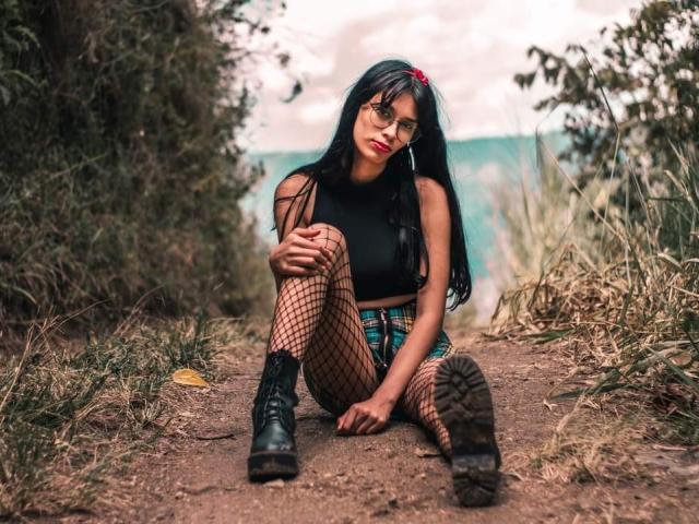 NycoleAngels with long black hair, glasses, red hairclip, black crop top, fishnet pants, plaid skirt, black boots, sitting on dirt path, hands on knees, looking at camera.