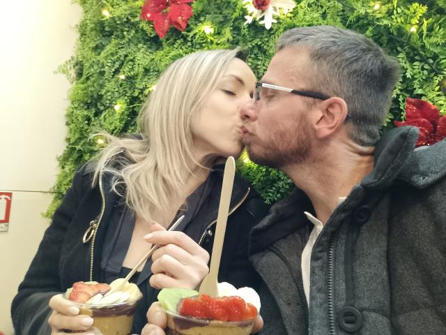 Annic with blonde hair and Annic with gray hair, beard, and glasses kissing while holding desserts with straws. Annic's tits are slightly visible under a black jacket.