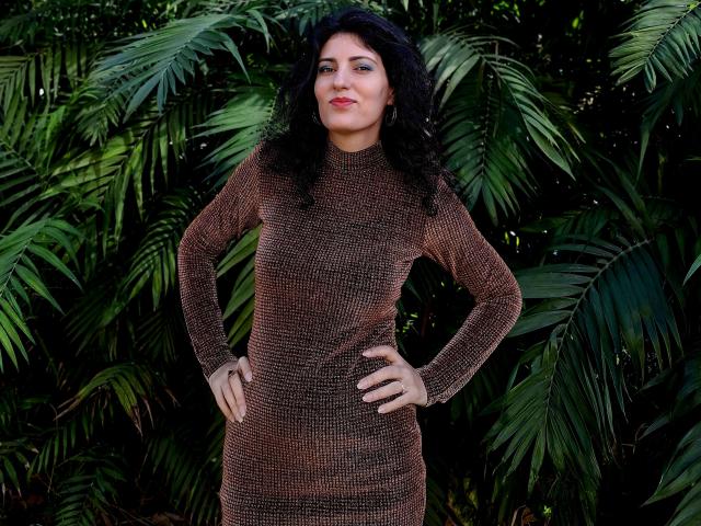 AddictedToUTS with curly black hair, red lipstick, and a brown, textured, long-sleeve dress, standing with hands on hips, in front of green, leafy foliage.