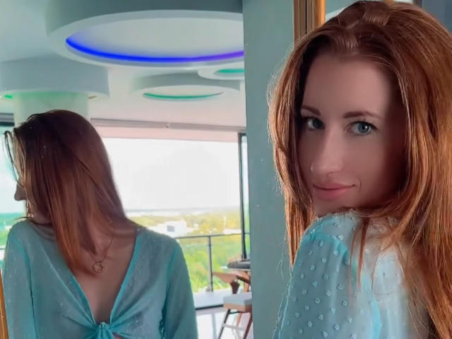 TheTyQueen, con cabello rojo, una blusa azul transparente y una sonrisa encantadora, mirando por encima del hombro, mostrando pequeños senos en el reflejo del espejo, en una habitación moderna y soleada..