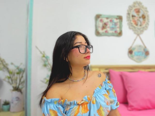 Genesiss, con cabello negro, gafas, lápiz labial rojo, vestido azul con hojas amarillas, un pequeño tatuaje en el hombro derecho, un collar, un cojín rosa y un trasero muy lindo..