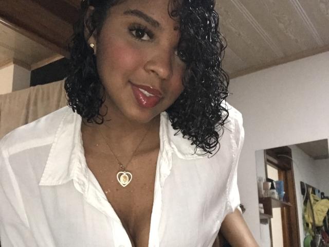 LisaMendez with curly black hair, wearing a white shirt, showing a heart necklace, has shiny red lips and a cute smile, her small tits slightly visible, in a cozy room.
