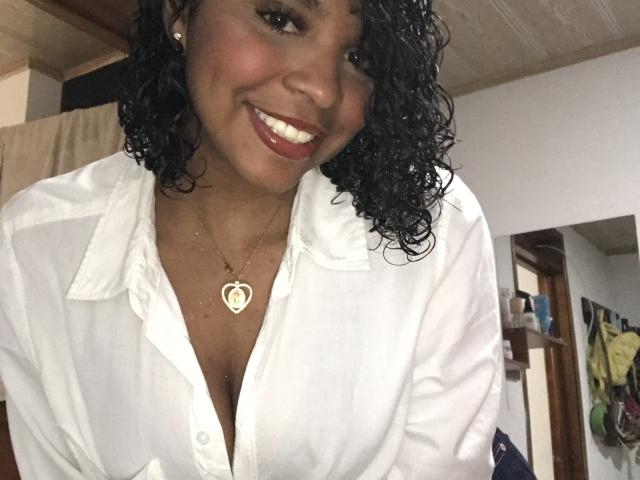 LisaMendez, sorrindo com cabelos pretos encaracolados, vestindo uma camisa branca de botões, mostrando o colo, usando um colar com formato de coração e batom vermelho, em um quarto de madeira..