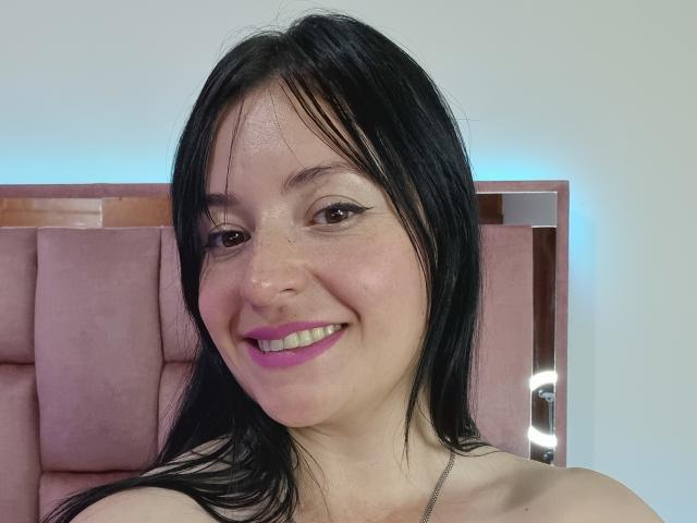 VaaleryStark with black hair, pink lipstick, and white skin, smiling at the camera, showing freckles and wearing a silver necklace, with pink upholstered headrest behind.