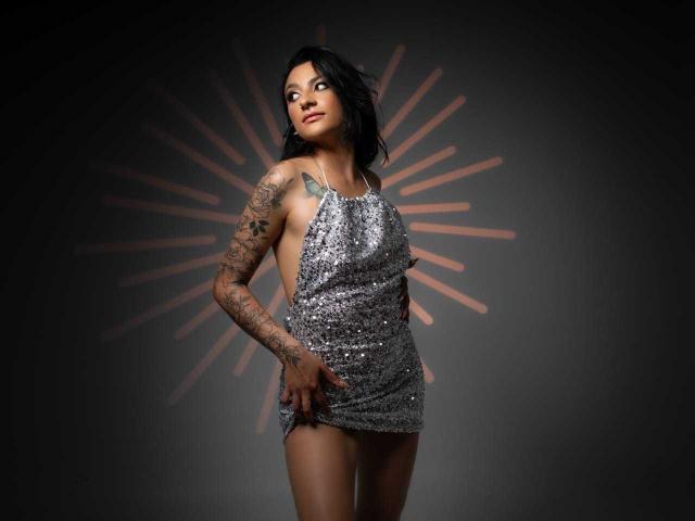 Alizep with dark hair, wearing a sparkly silver halter dress, tattooed arm, and hand on her ass, standing confidently, looking up with a slight smile.