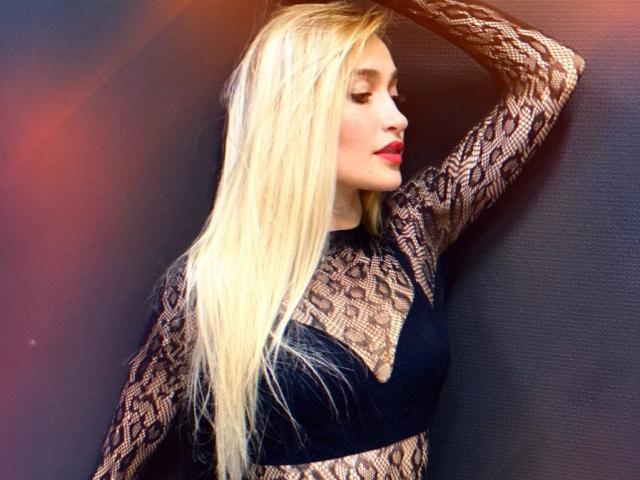 KateOweens with long blonde hair, red lipstick, and black lace top, hand in hair, showing off her tits and lace pattern, standing against a dark textured wall.