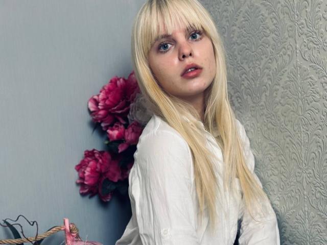 Lialit tiene el cabello rubio largo, ojos azules y piel pálida, lleva una camisa blanca, con flores rosas detrás, mirando ligeramente hacia un lado con una expresión neutra..
