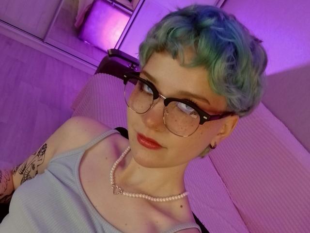 ShellyLove69 with green curly hair, glasses, red lipstick, white tank top, pearl necklace, tattoos on arm, purple lighting, looking at camera, sitting on bed.