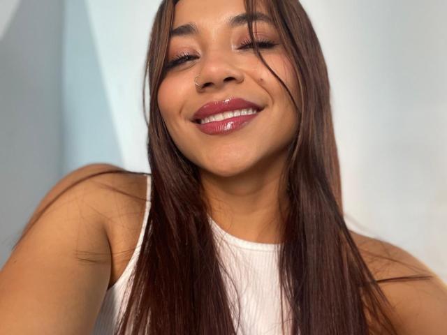 Sophie Denisee met glanzende, roze lippen, bruin, steil haar, een wit tanktopje, met gesloten ogen en een glimlach, bruine huid, een schattig gezicht en een kleine neusring, ziet er gelukkig en ontspannen uit..